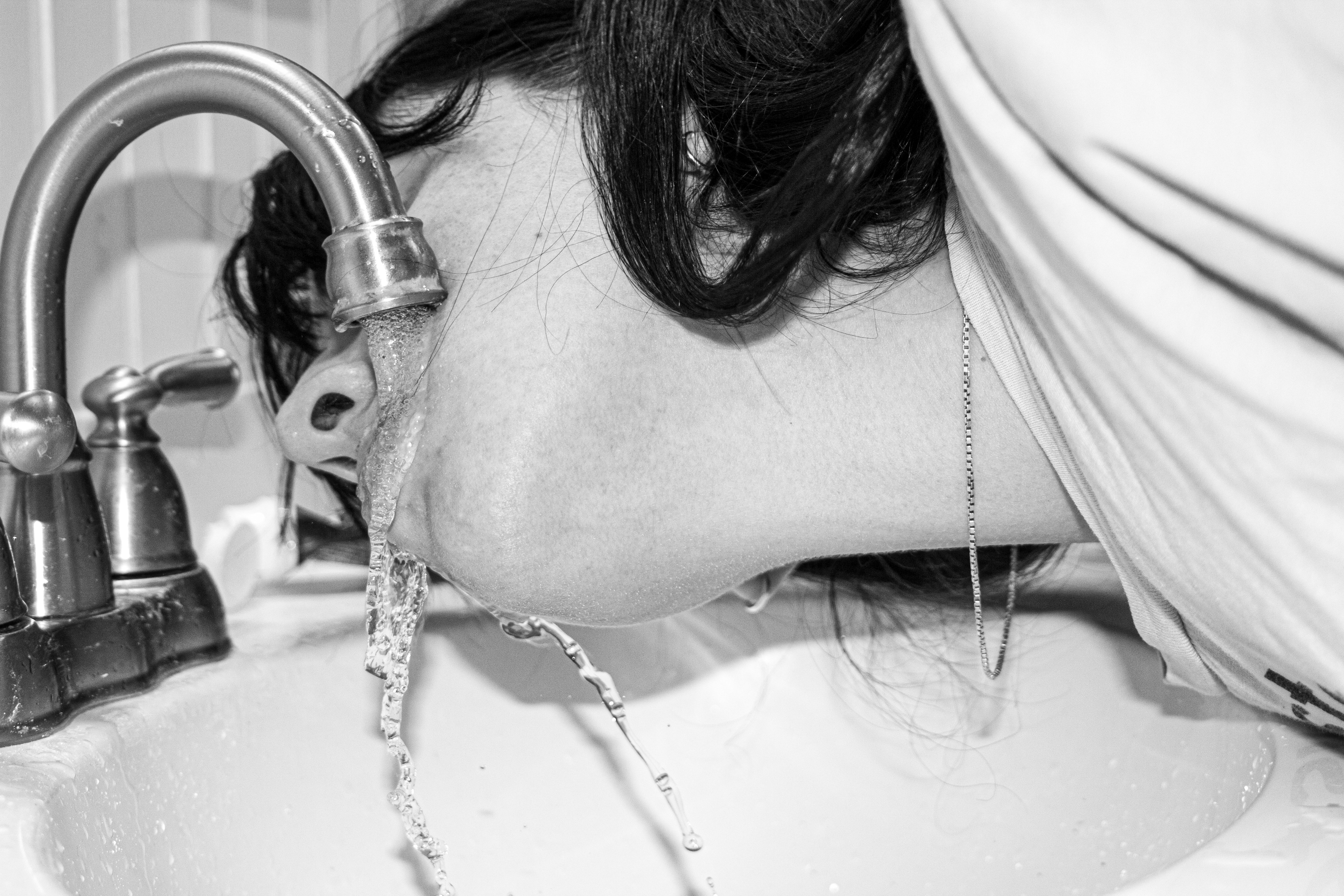 A woman drinks out of a sink, with water straming down. It is in black and white and you can see her chain. 