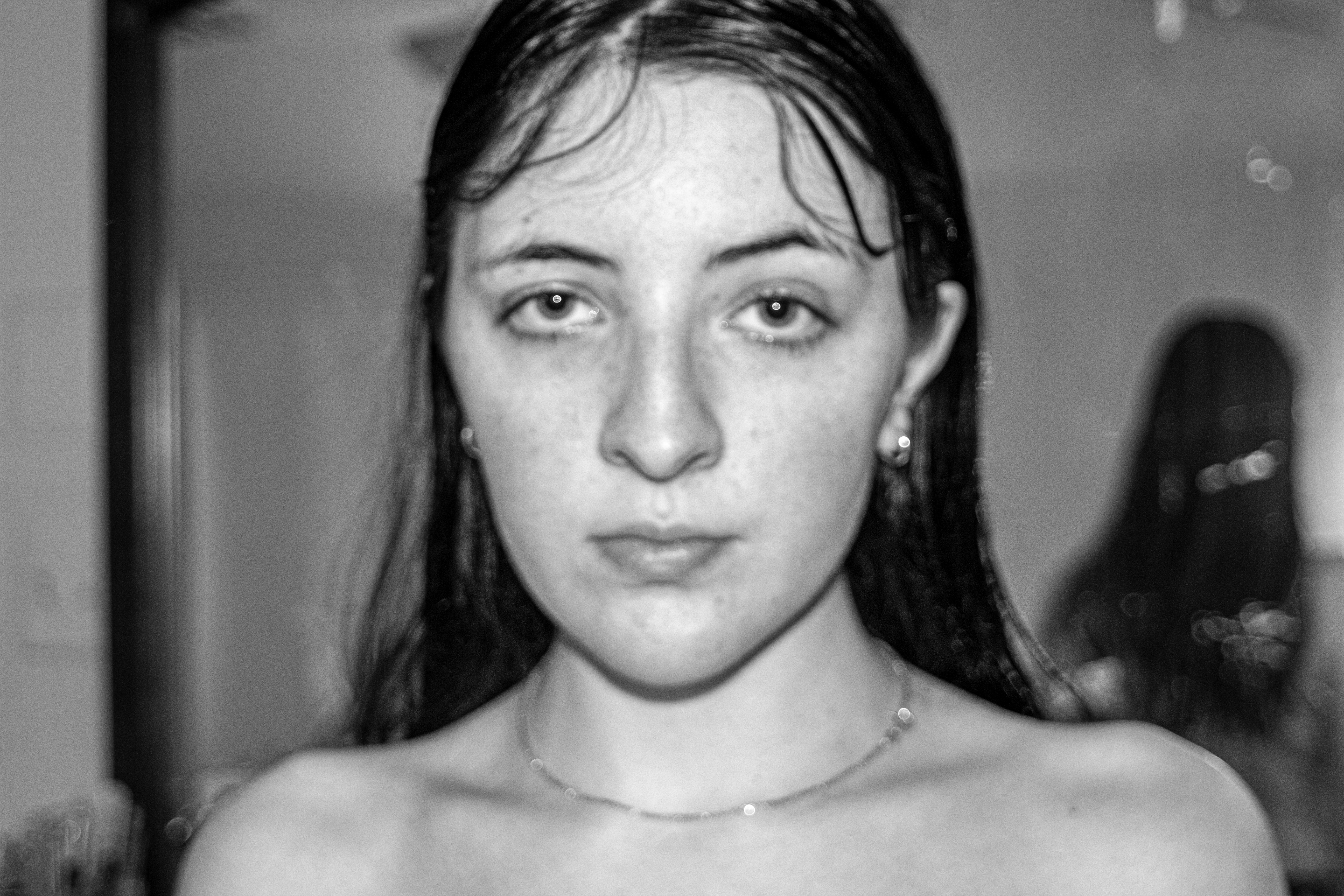 A woman without a shirt looks directly at the camera with a look of complex unrest. A naked version of the woman is seen, blurry in the mirror in the background with her back to the woman.