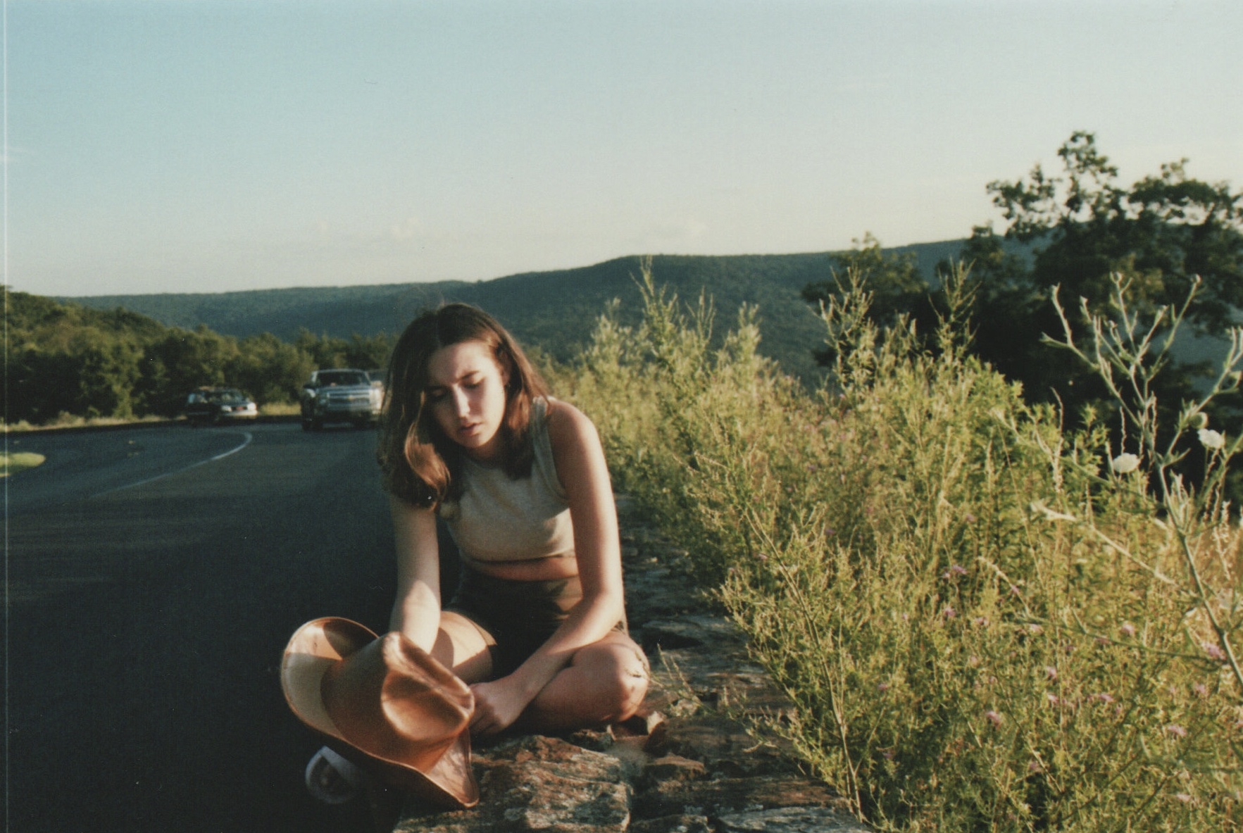 A woman sits on a scenic overlook, looking tired. She is holding a cowgirl hat and the main color in the photo is green.