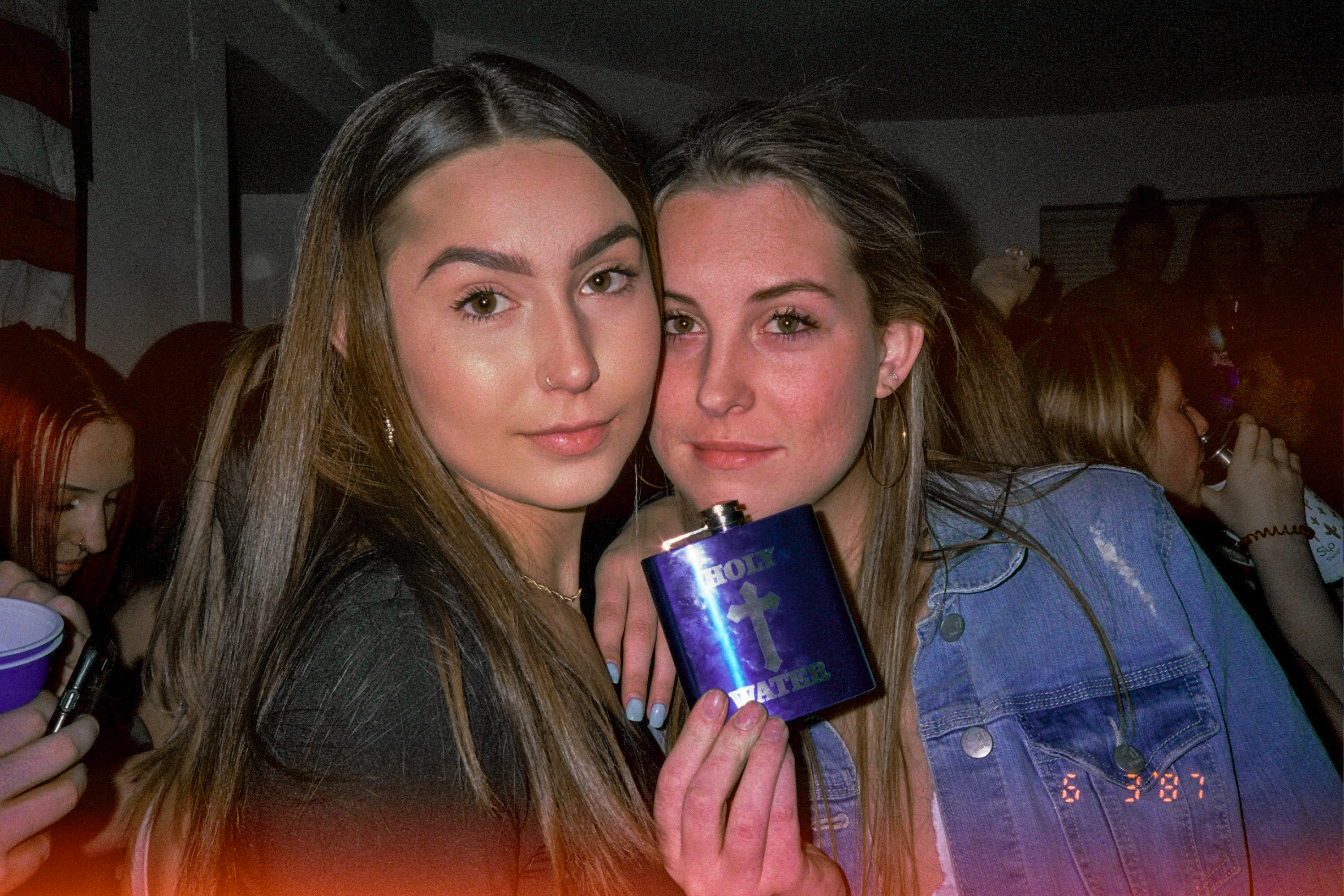Two women are at a party and are looking into the camera. The woman on the left is wearing a jean jacket and is holdind a flask that says holy water. 