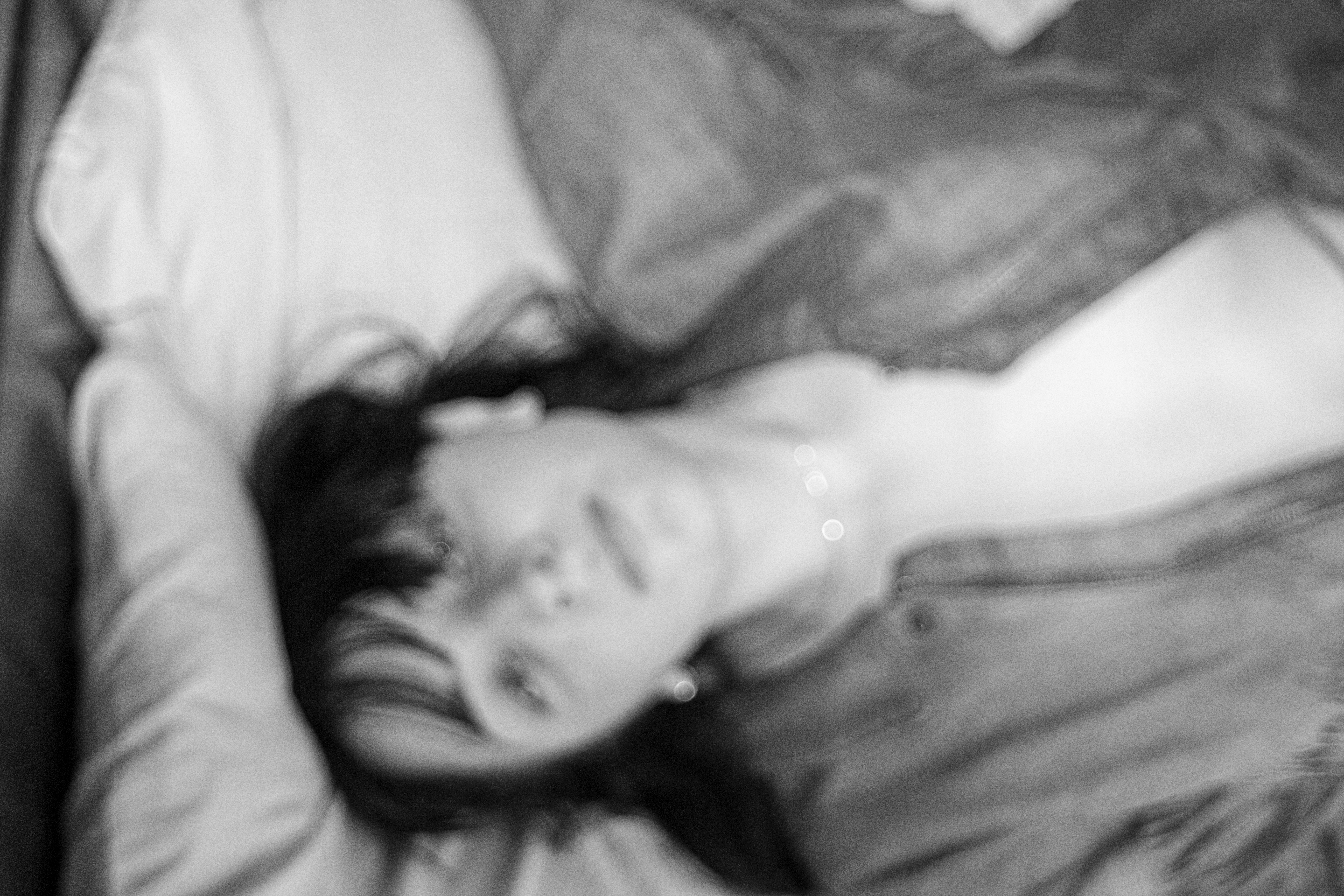 A woman on a bed looks up at the camera. She has dark hair and is wearing an open jacket and no shirt underneath.