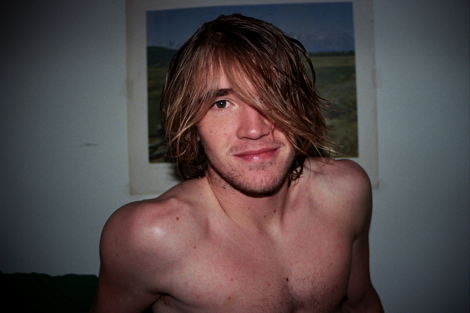 A man looks into the camera not wearing a shirt. There is a blue and green poster of a landscape hung on the wall behind him.