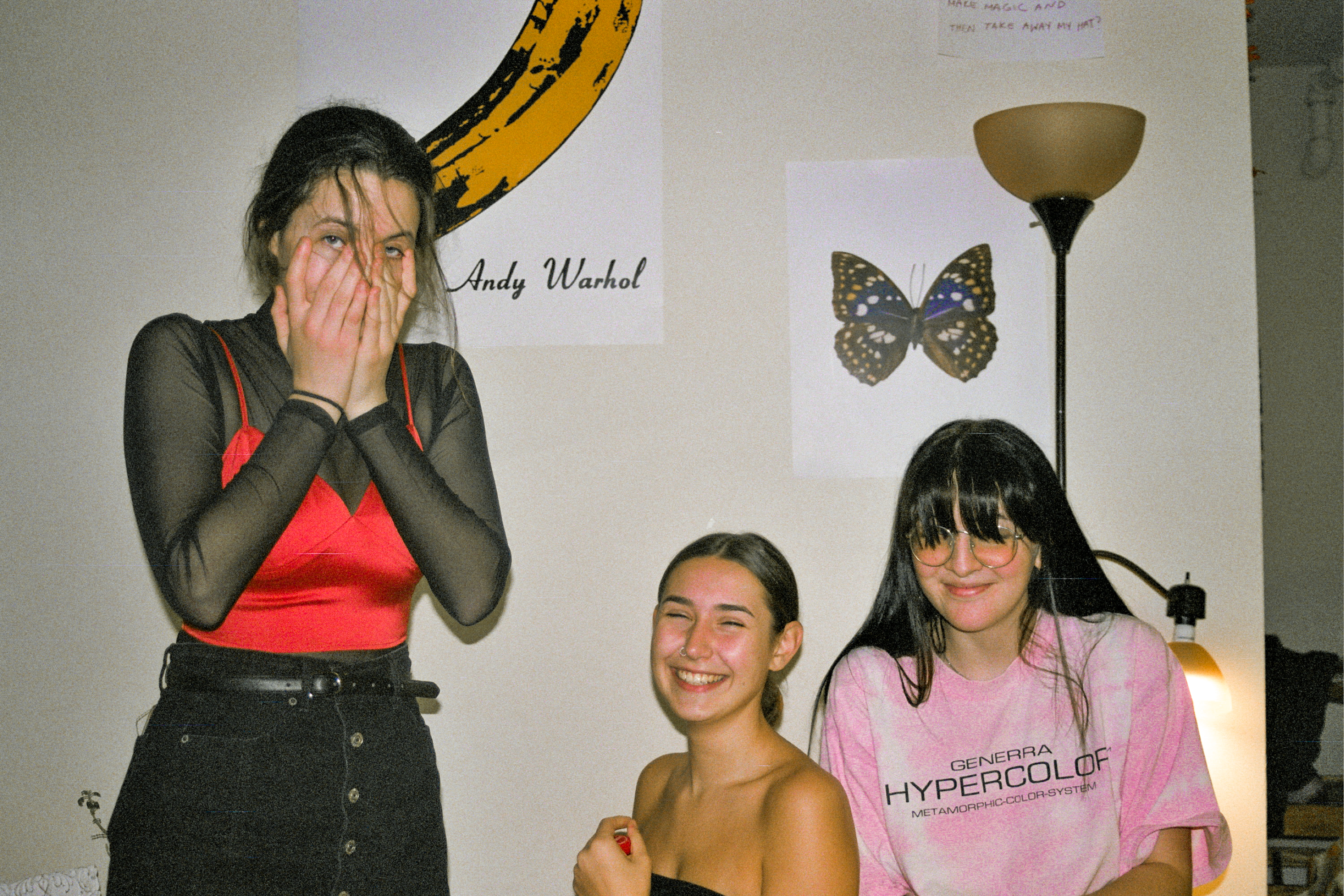 Three girls at different levels look into the camera. There is a velvet underground poster in the back and a butterfly poster and the girls are all laughing in different ways.