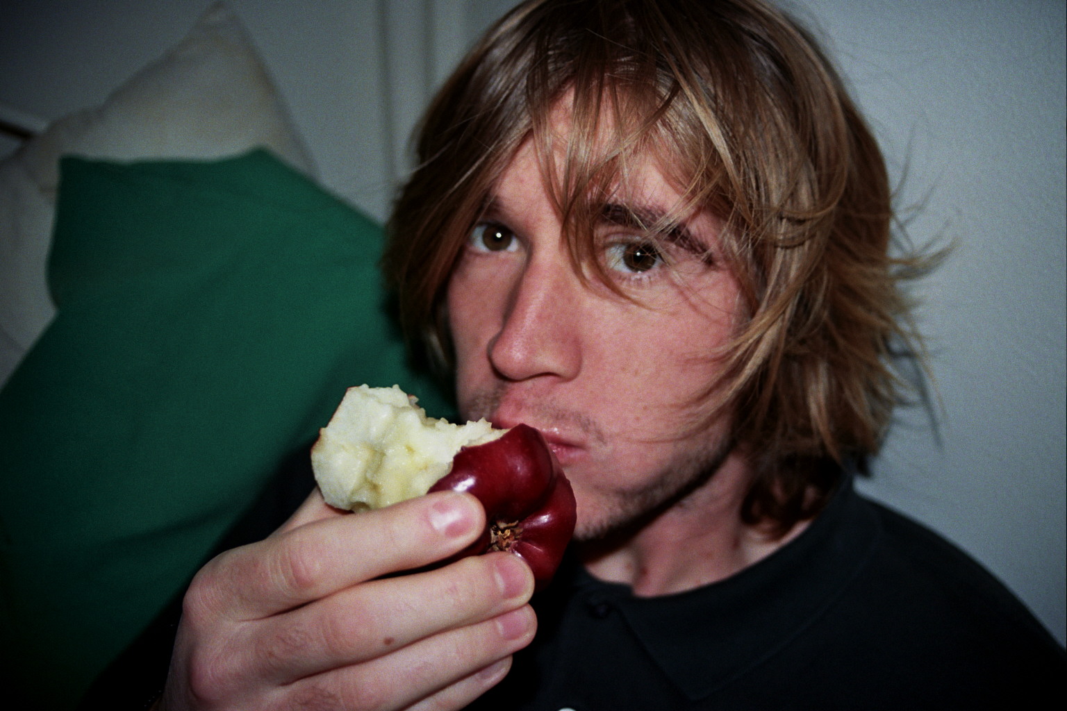 A man is shot in color with flash eating an apple. He is sitting on a bed with a green pillow behind him and has long blond hair.