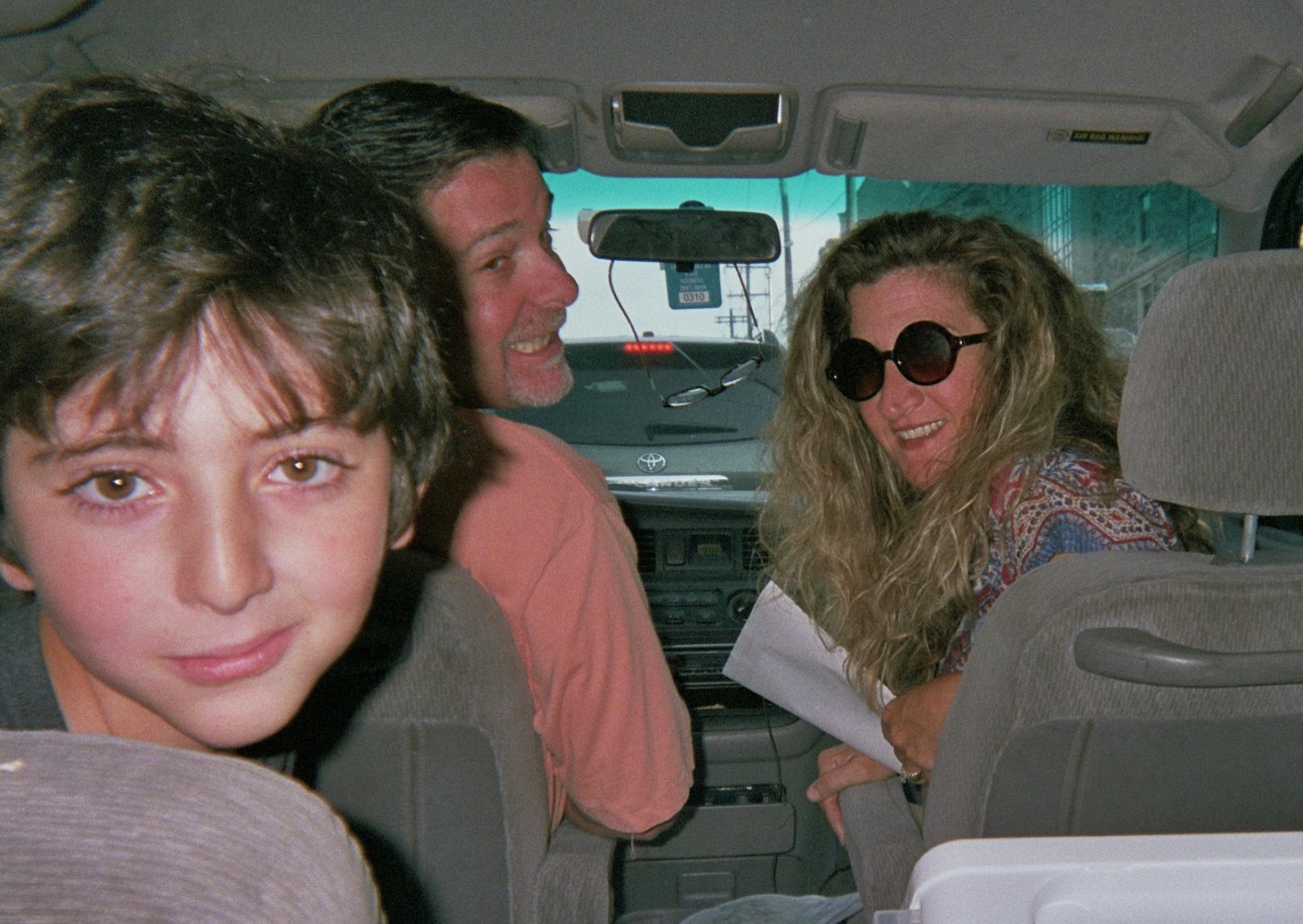 A family, a mom, a dad and a little boy are in a car. They are all turning their heads backwards to the person taking the photo. 