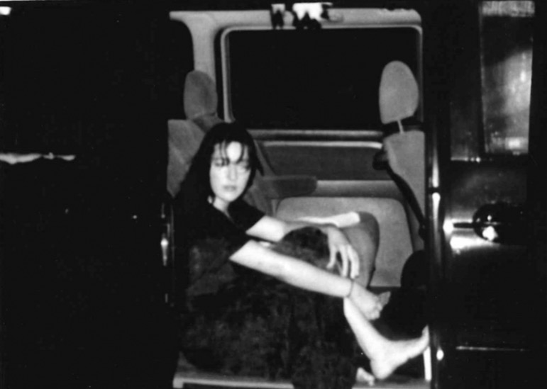 A girl sits in an open car in the rain at night. The photo is blurry and in black and white,.