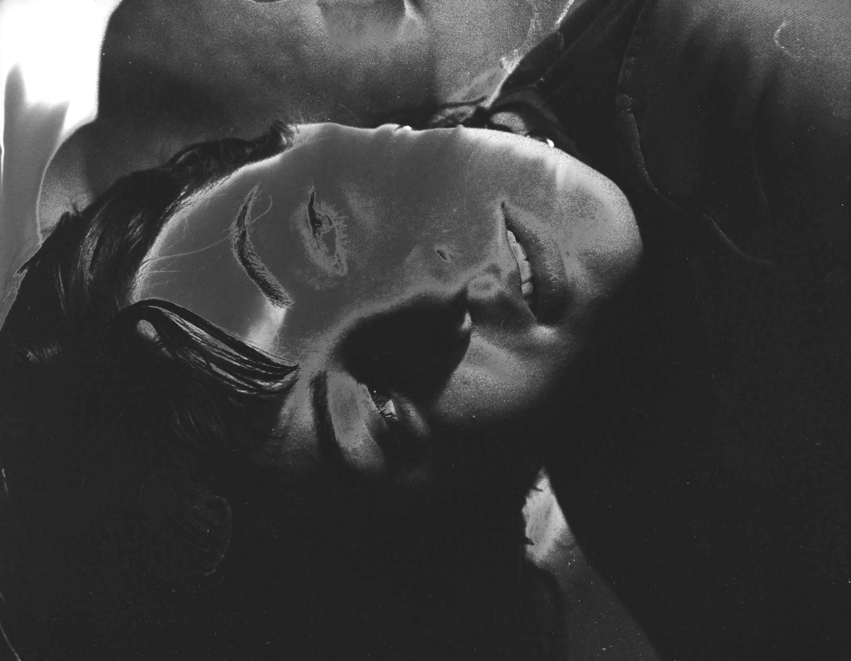 A picture of a woman bent over in black and white that is solarized so it is mostly black. You can only see her face and a strange organic background in the back and the photo is moderately to very close on the face.