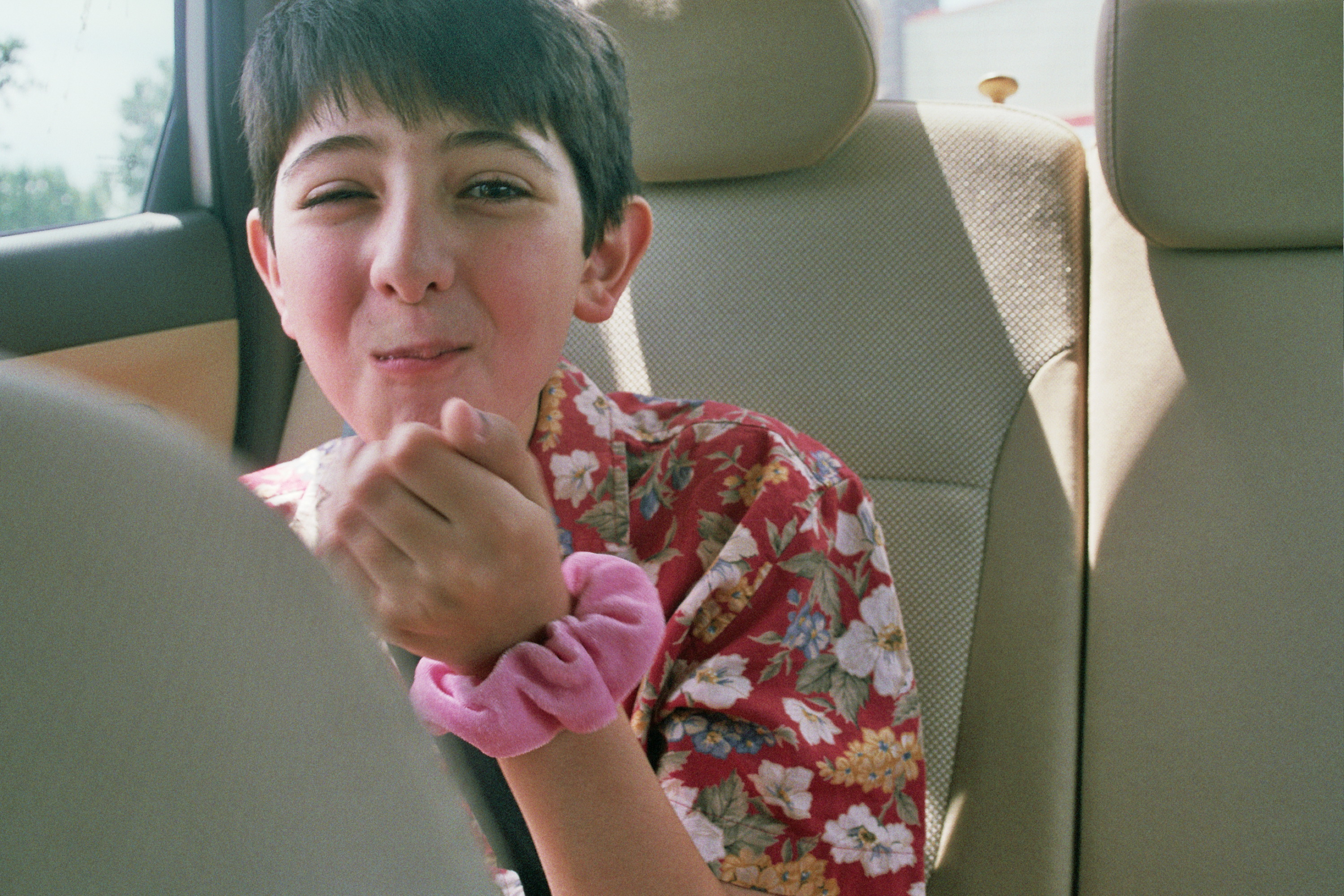 A little boy sits in the backseat with something in his mouth. He is laughing and has a pink scrunchie around his wrist.