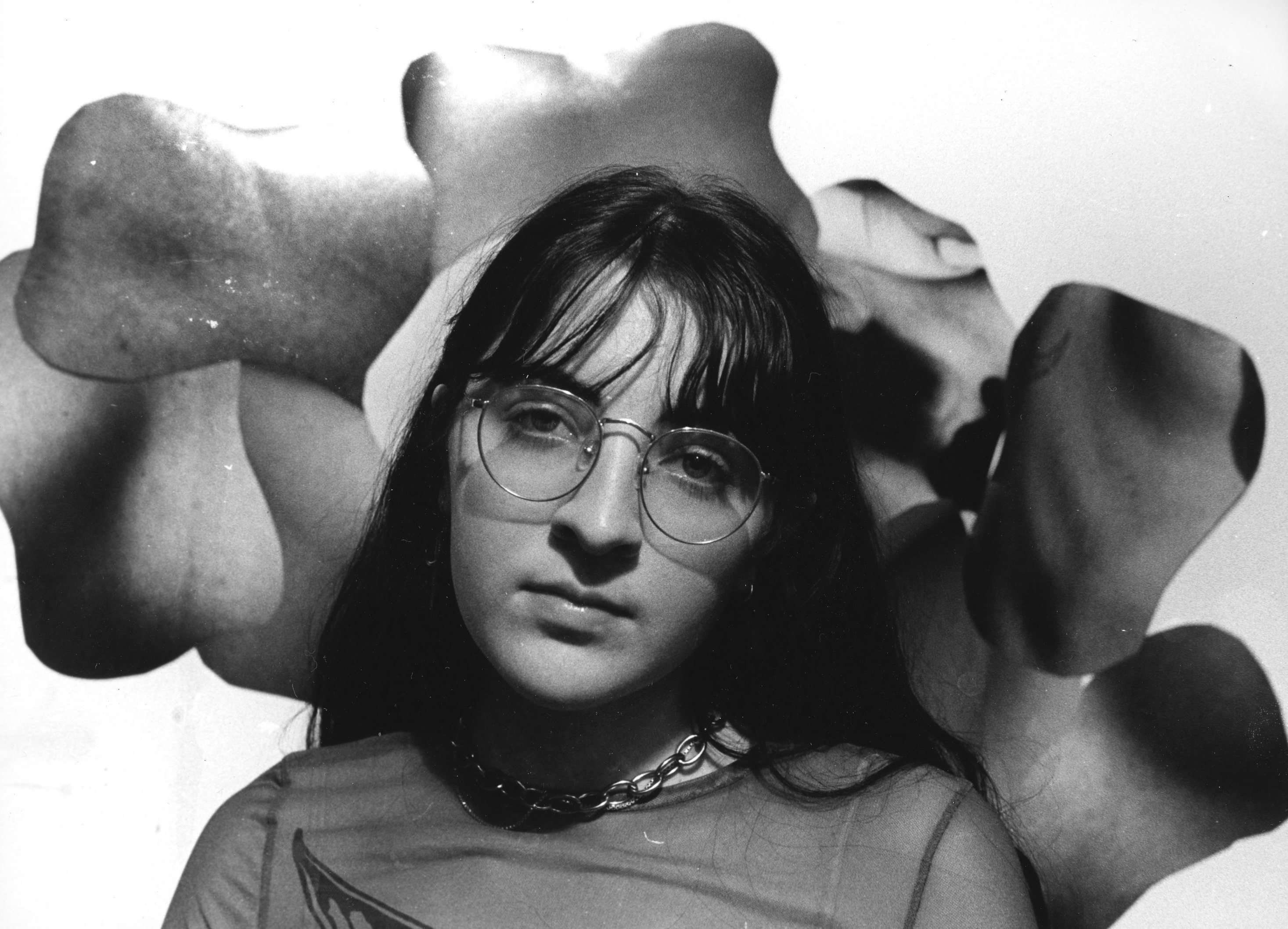 A woman sits in front of a strange collage of body parts.  She is sitting directly in the ceneter and is wearing a mesh shirt and glasses. The image is in black and white.