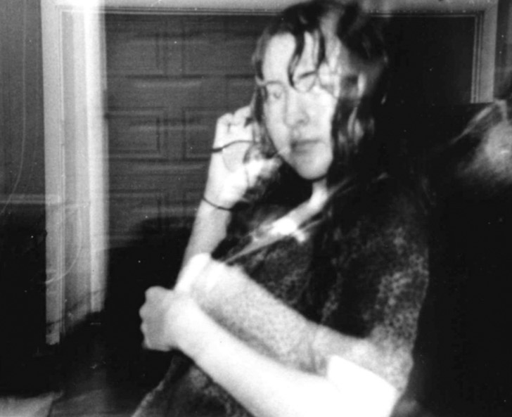A girl leans back on a car in the rain. She is holding her glasses and the photo is blurry and in black and white.
