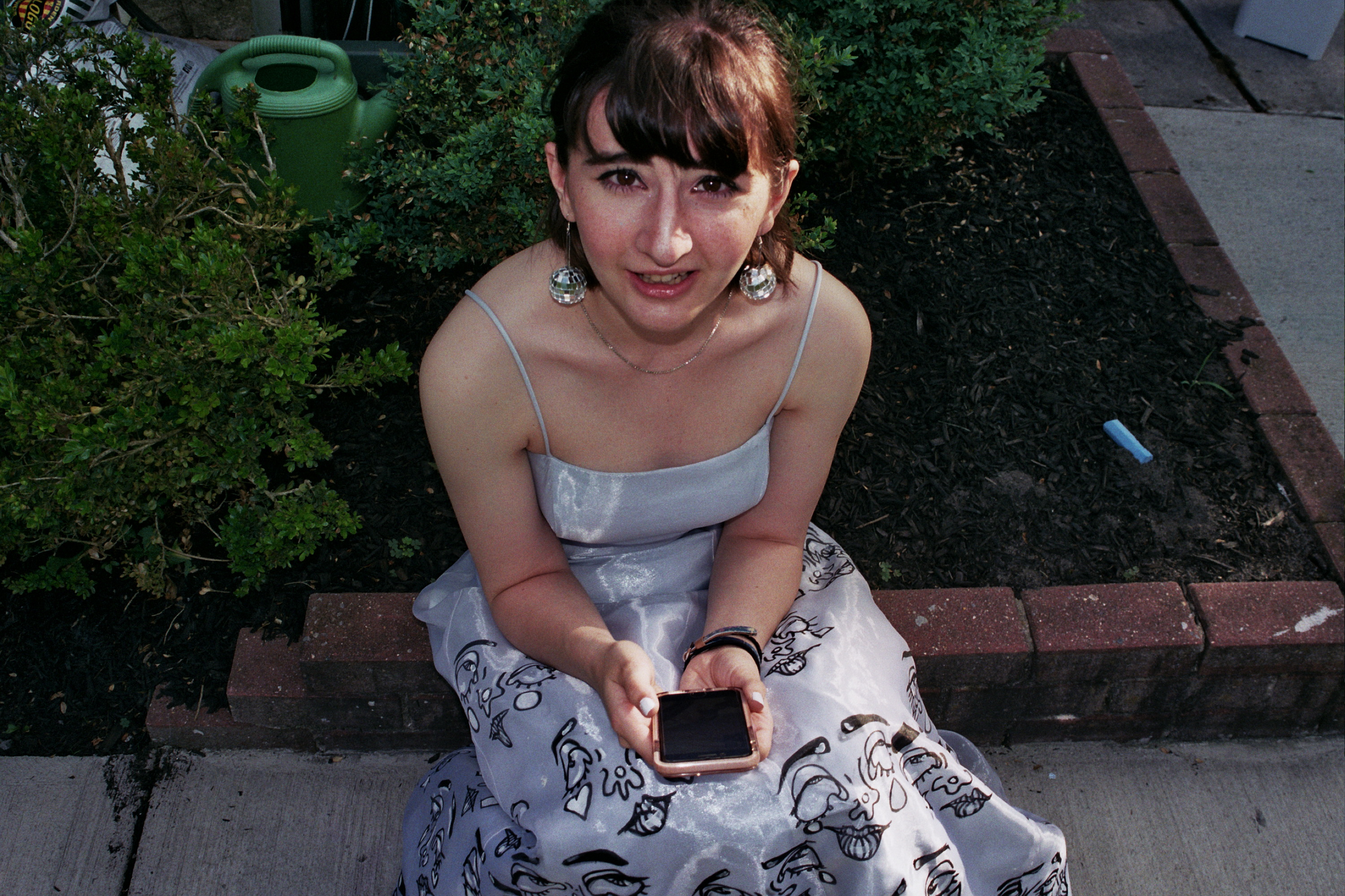 A woman sits on an outdoor setting wearing a blue painted prom dress. She looks up inot the camera 