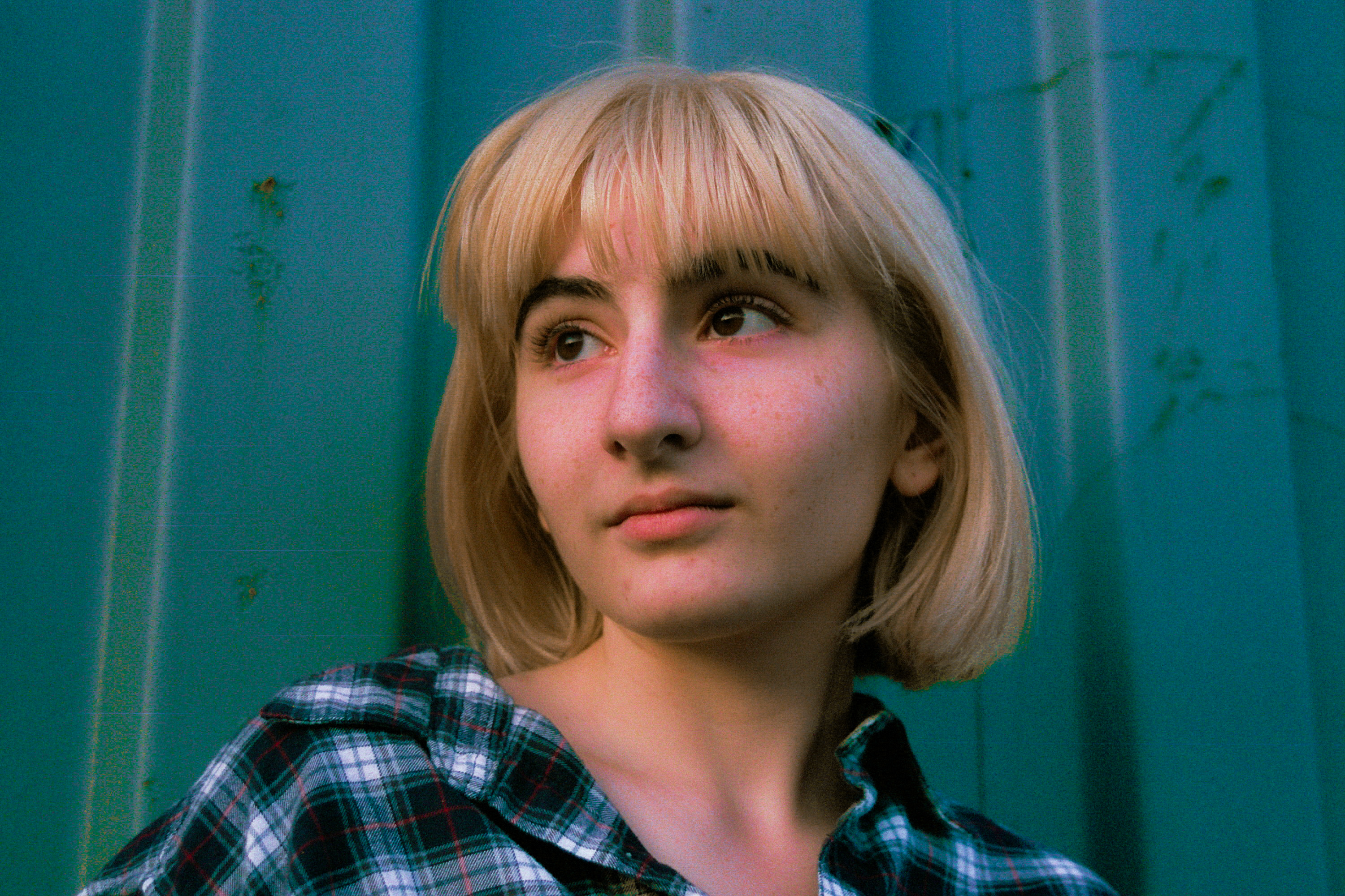 A woman with short blonde hair looks to the left.  The photo is in color and the background is blue and she's wearing a flannel shirt.
