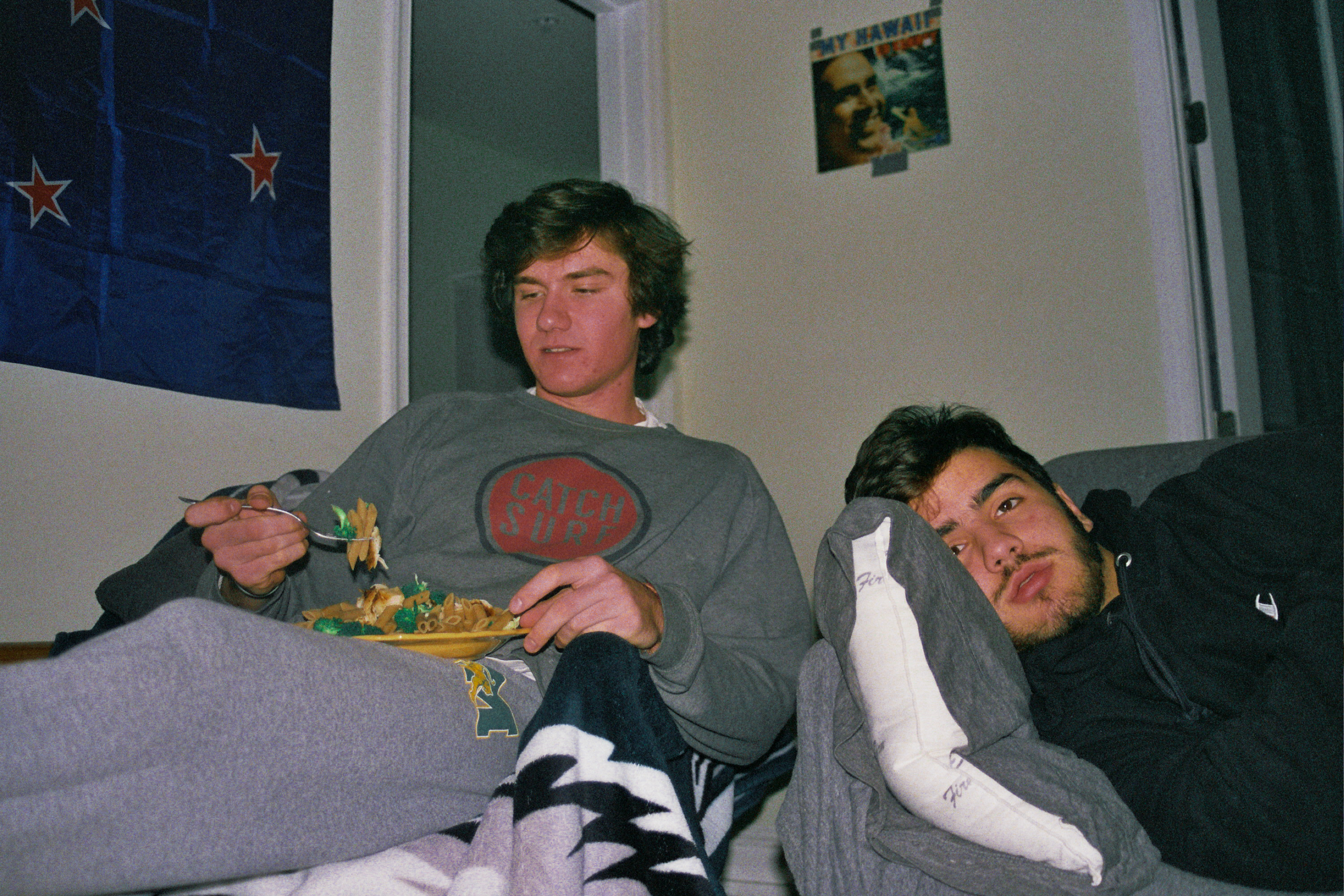 Two men sitting in a dorm room, one sitting up the other laying down. One is eating dinner and the other is looking right in the camera. 