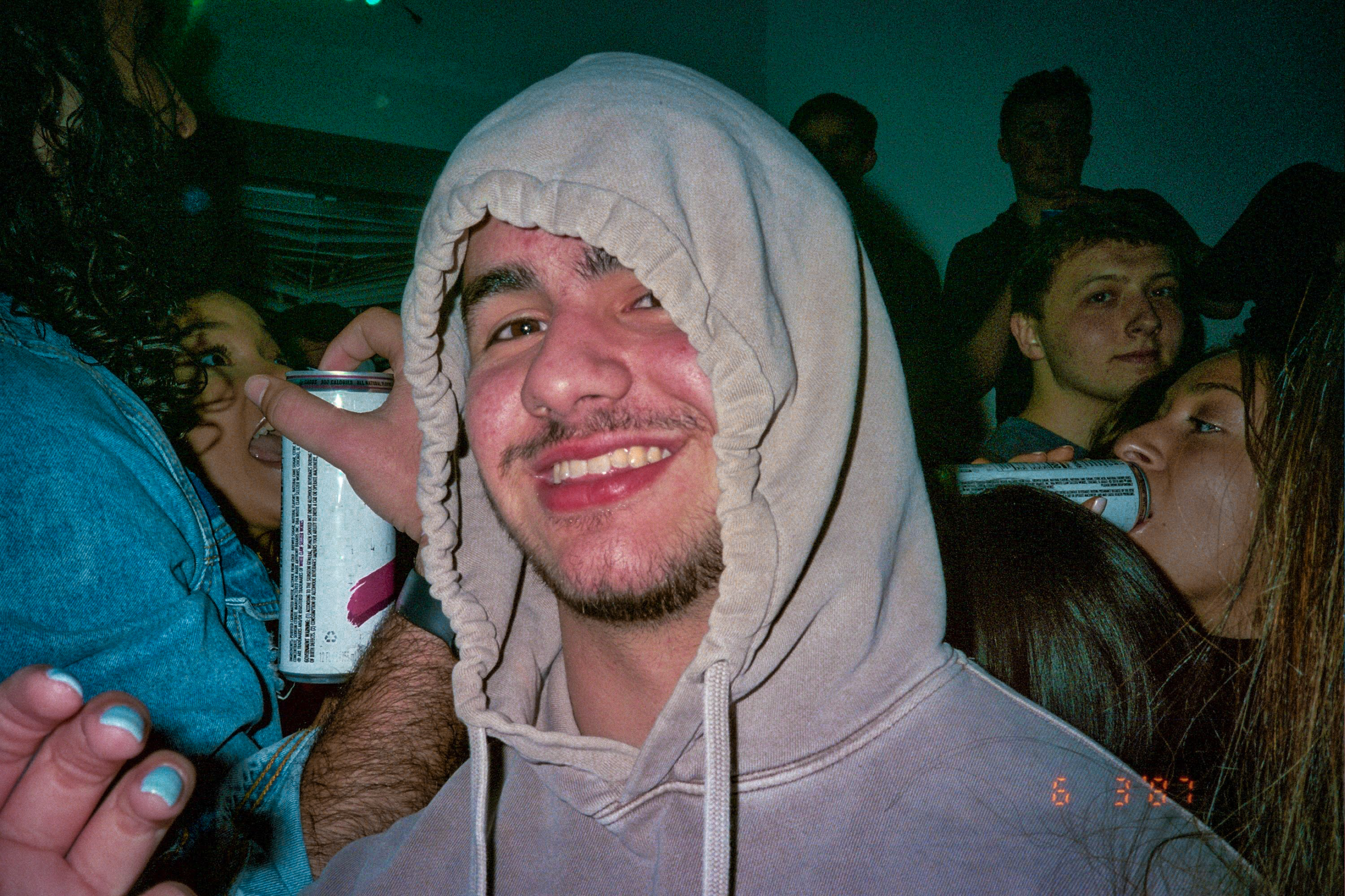 A color photograph of a man with a beard and mustache at a party. He is wearing a gray hoodie and the people behind him are in levels and drinking. 