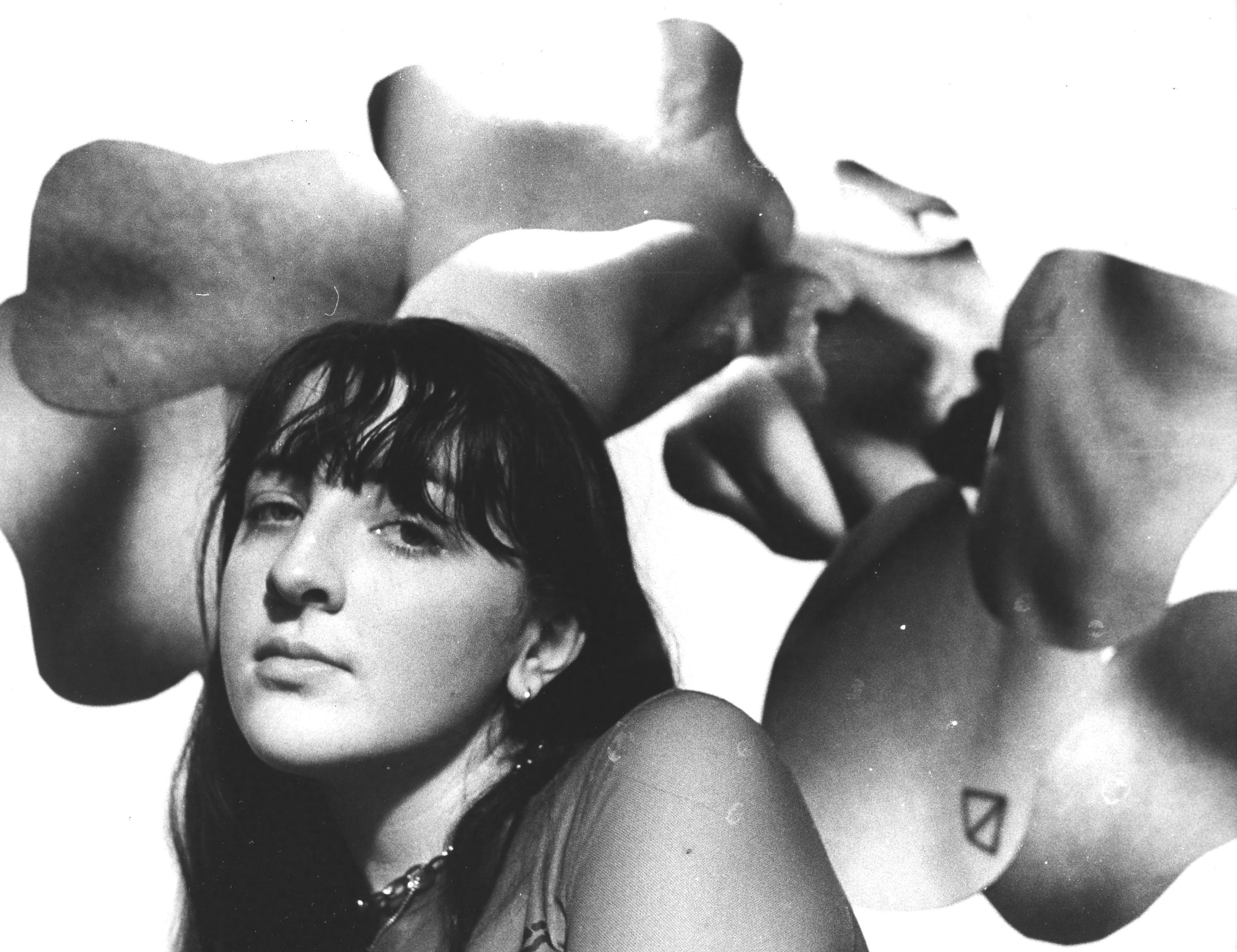 A woman sits in front of a strange collage of body parts.  She is titled slightly and is wearing a mesh shirt. The image is in black and white.