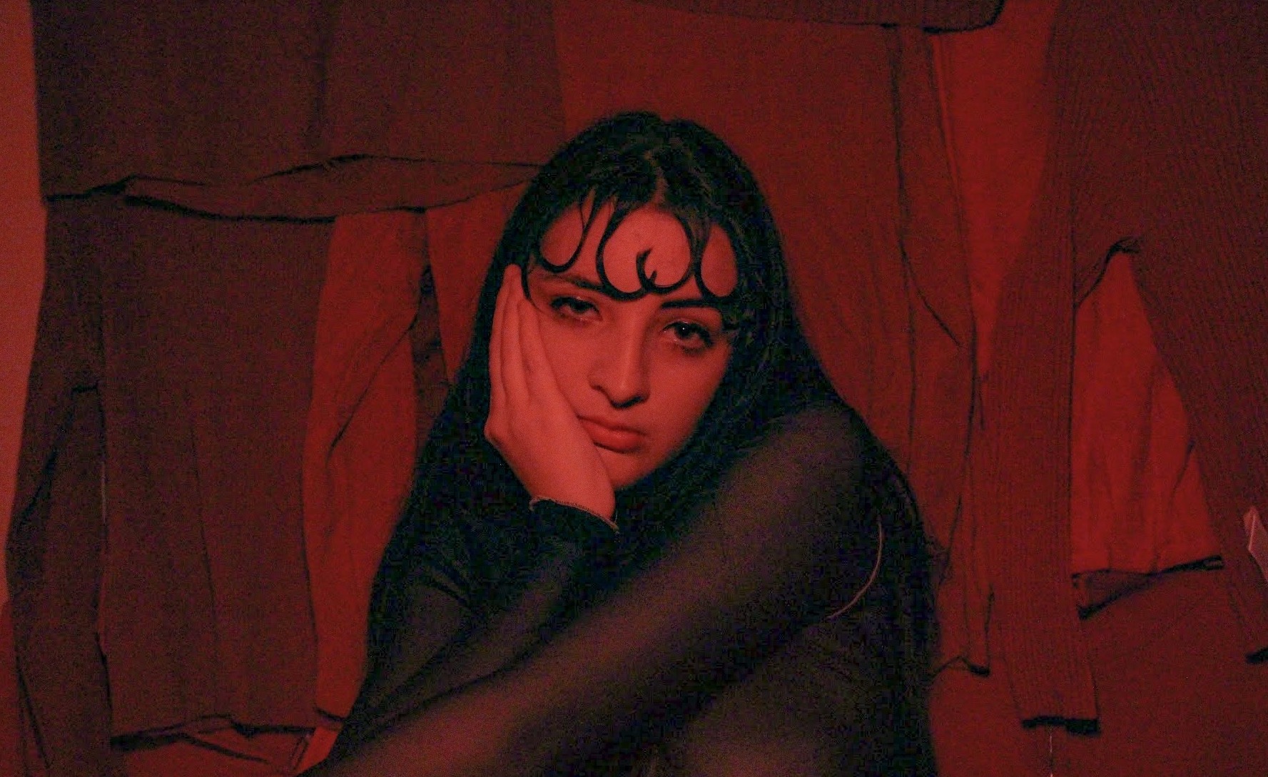 A woman in all red with a red background looking at the camera with a hand on her face.