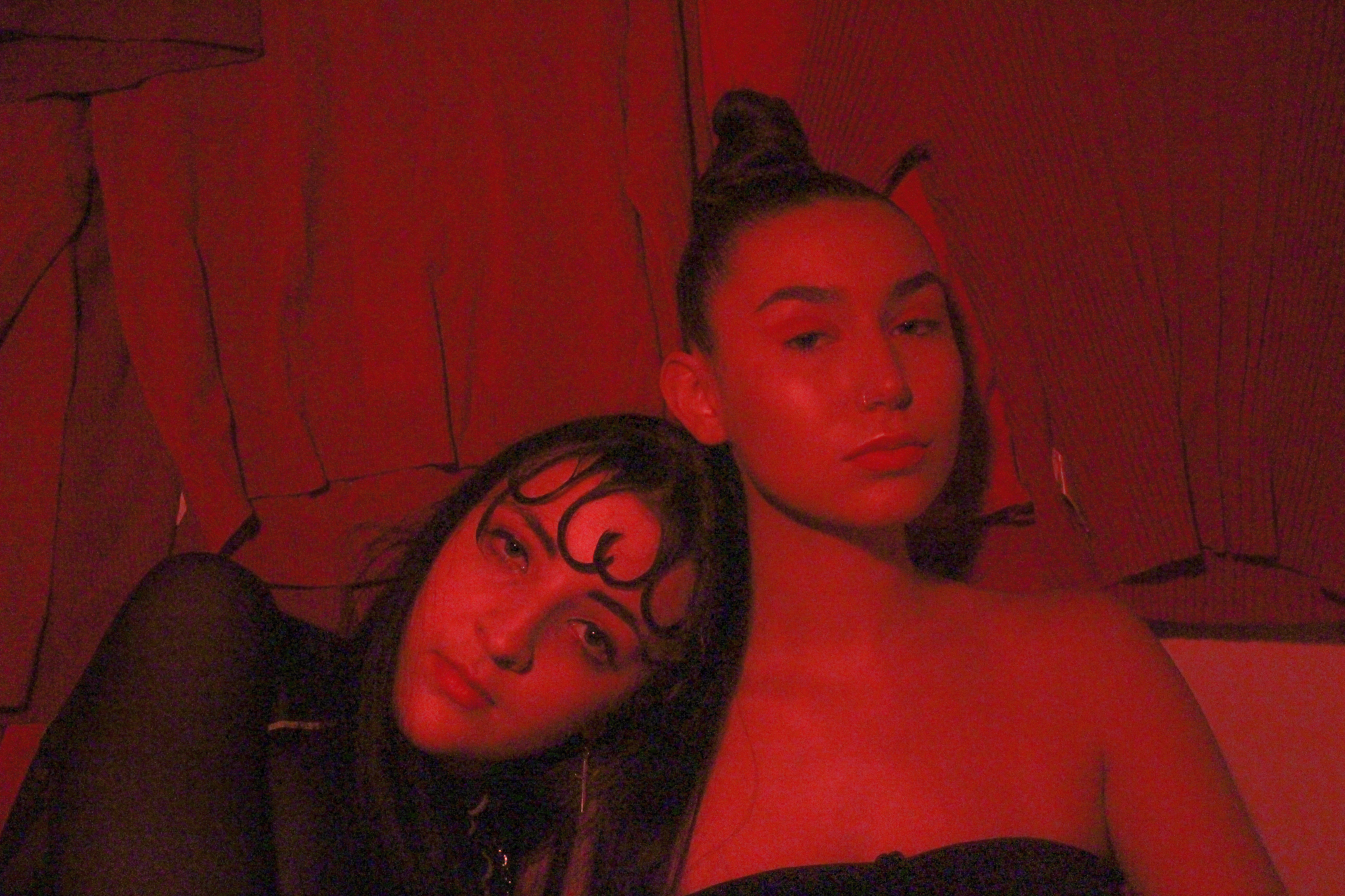 Two women lean on each other in an all red setting, one with her head on the other's shoulder. 