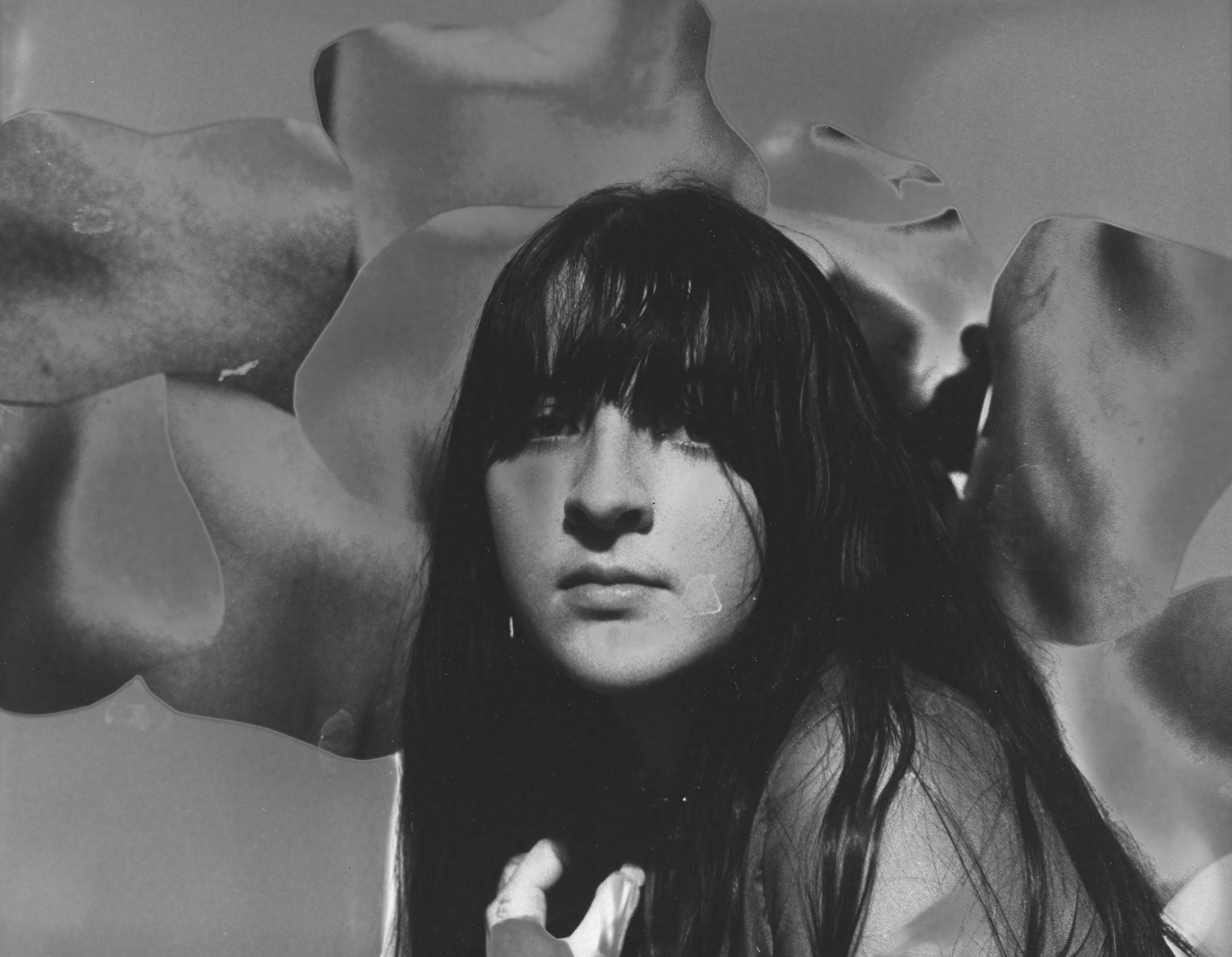 A woman with bangs is sitting in front of a strange organic background of body parts. Her shoulder is to the camera and she is looking in the camera nad the image is in black and white and solarized.