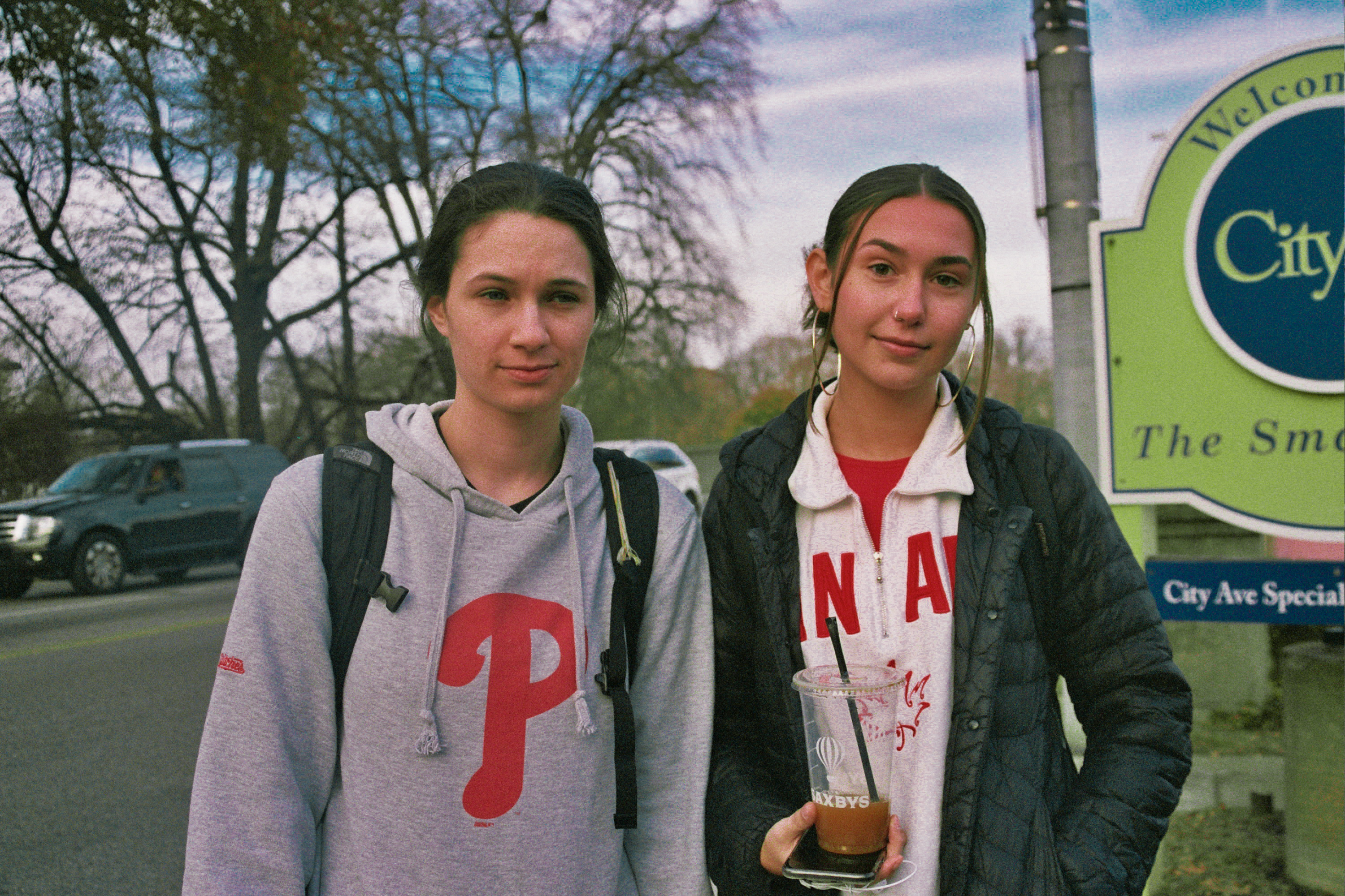 Two women standing next to each other next to a road. The woman on the right is holding an iced coffee and the woman on the left isn't looking at the camera and is wearing a shirt with the Philadelphia Phillies sweathsirt.