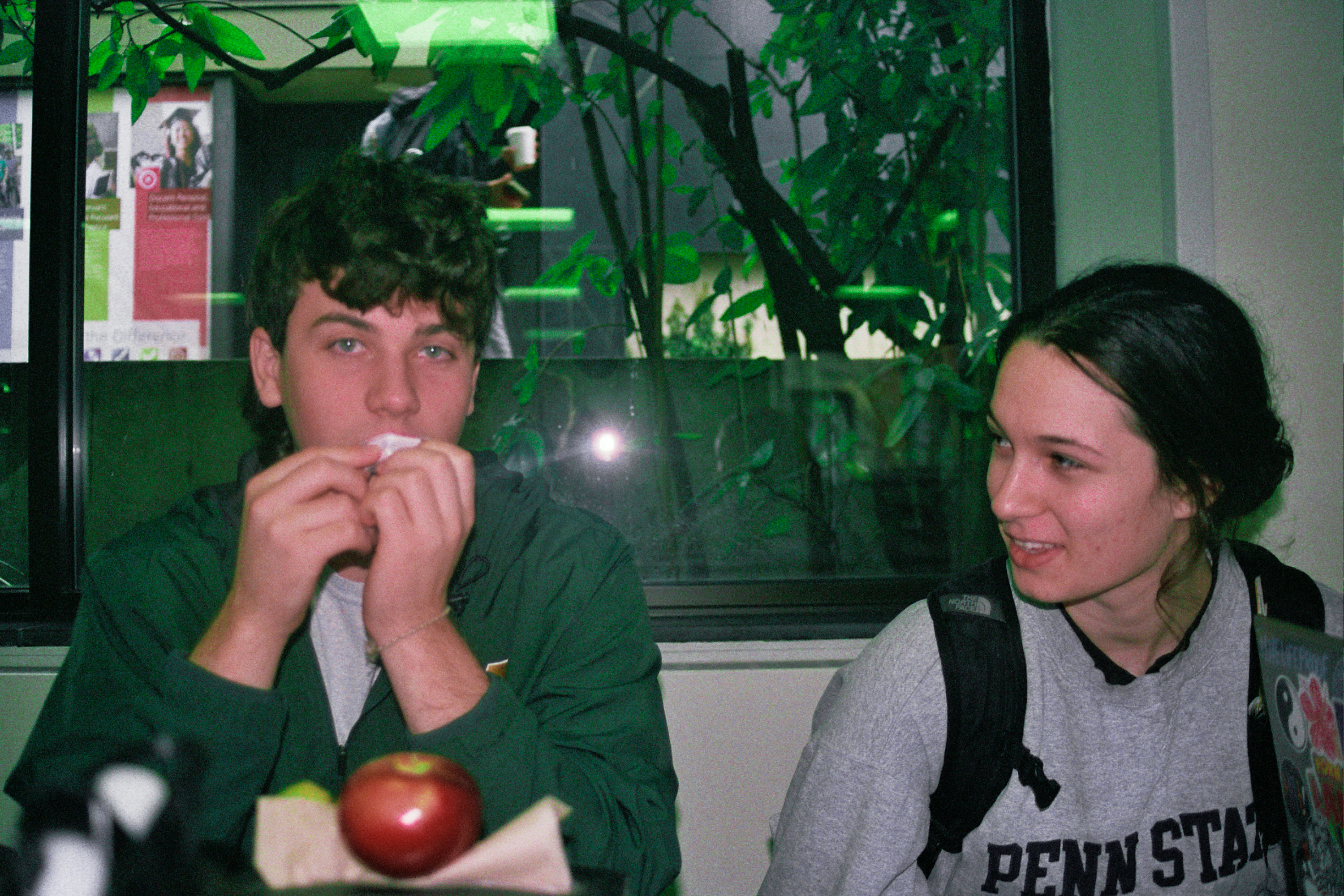 A color image of a a man on the left and a woman on the right with green in the background. The man is wiping his mouth with a napkin and the woman is looking at him. There is an apple in front of them.