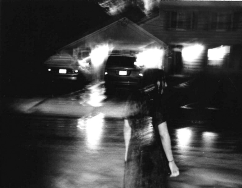 A girl wanders out into the street at night. The photo is rainy, blurry and in black and white.