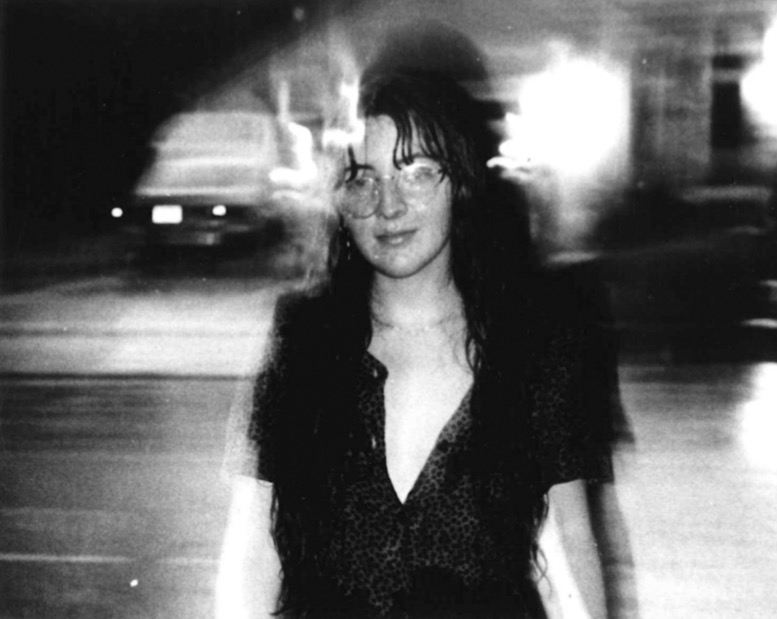 A woman stands in the street at night wearing a dress. The image is blurry and black and white and it is raining in the image.