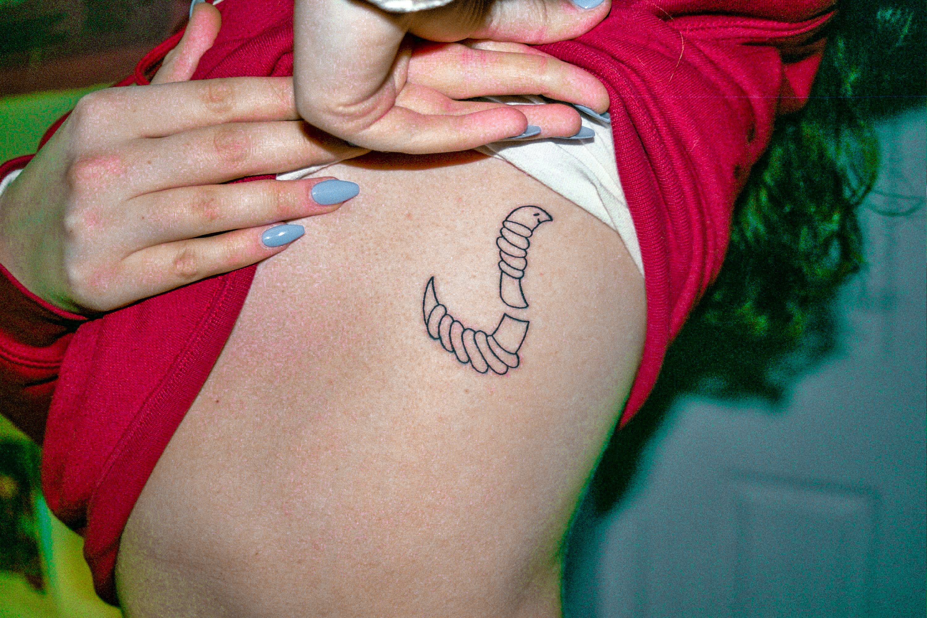 A woman in a red shirt is showing a tattoo of a little worm cut in half. The background is green and her fingernails are long and blue. 